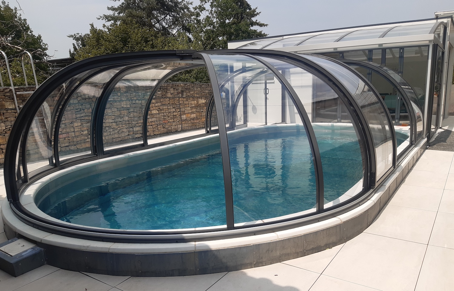 20260731_132509 Halbhohe, transparente Poolüberdachung über einem ovalen Außenpool mit Verbindung zu einer Terrasse und einem Wohnhaus.