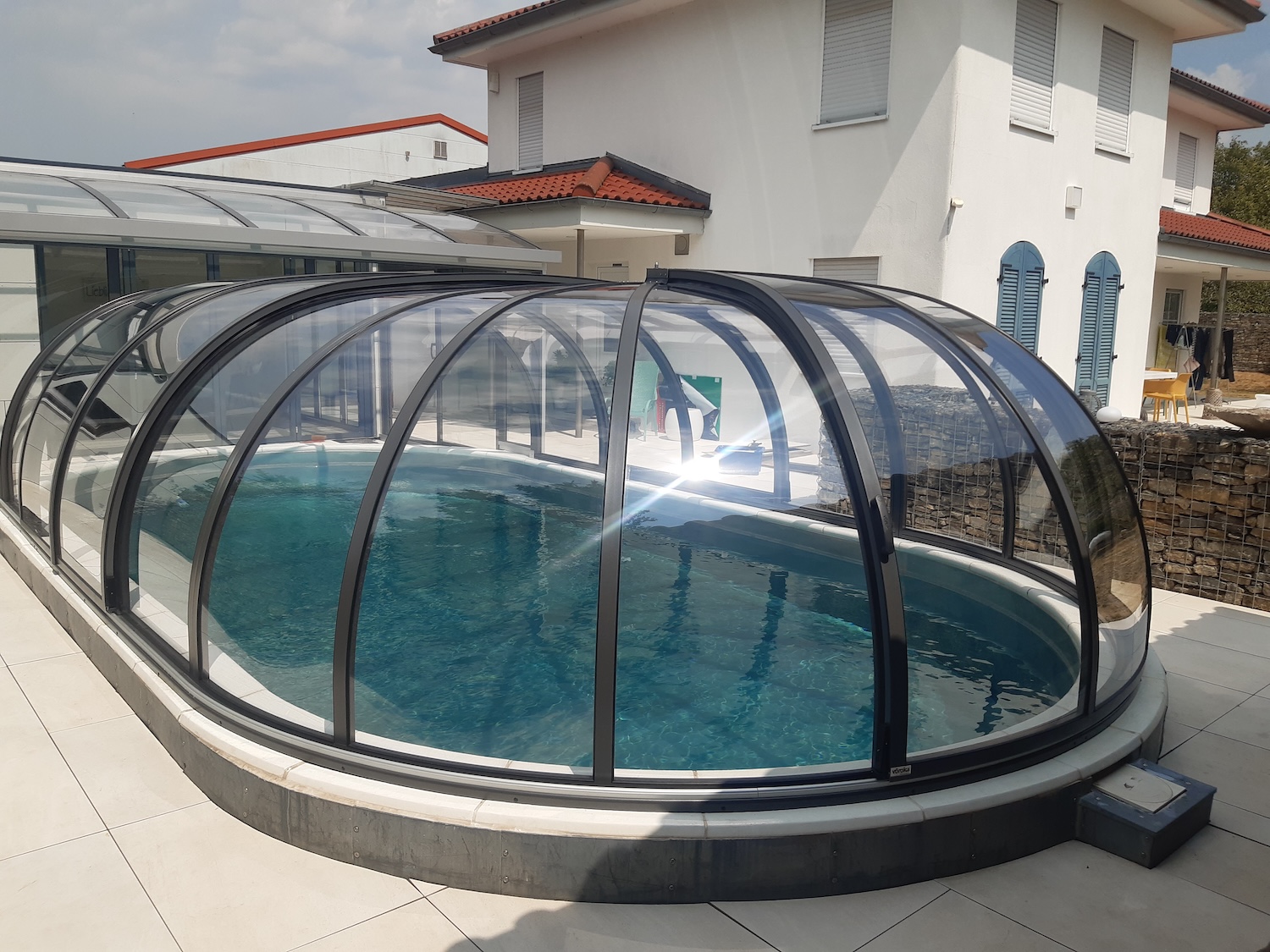 20260731_132650 Halbhohe, transparente Poolüberdachung über einem ovalen Außenpool mit Verbindung zur Terrasse und zum Wohnhaus.