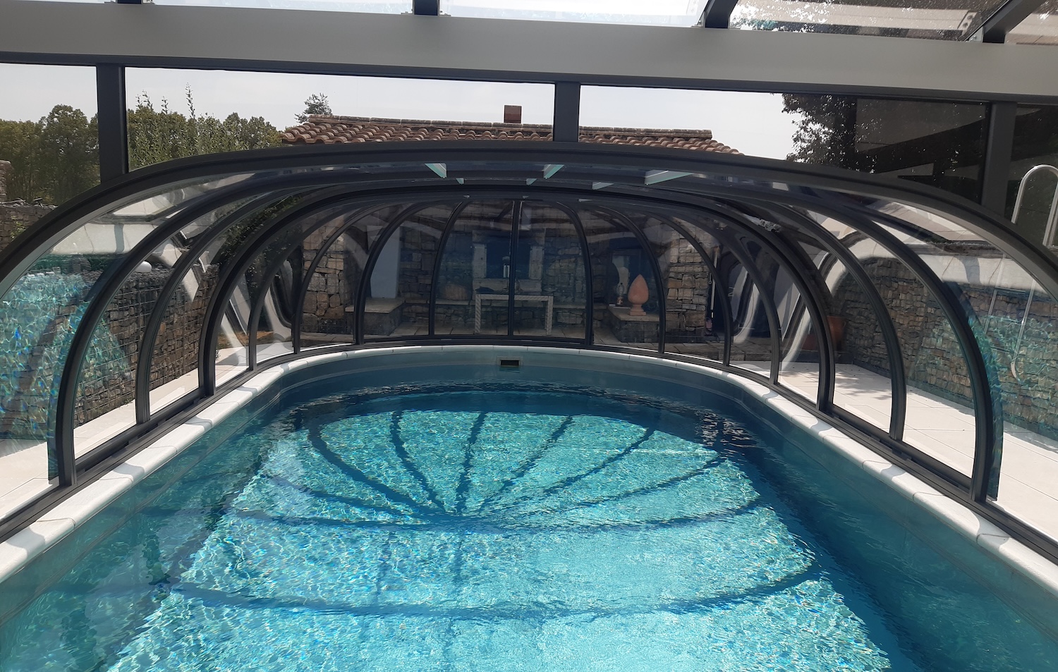 20260731_132746 Blick unter eine transparente Poolüberdachung auf einen geschützten Außenpool mit klarem Wasser.