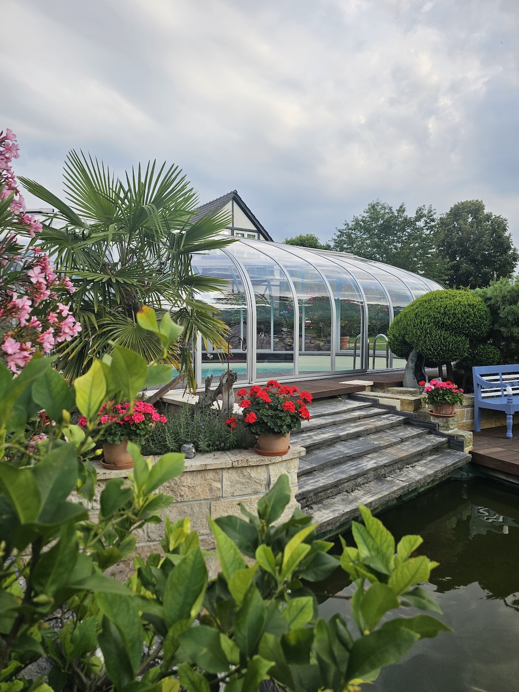 20260731_184507 Transparente Poolüberdachung in einem üppig bepflanzten Garten mit Palmen, Blumen und Steintreppe