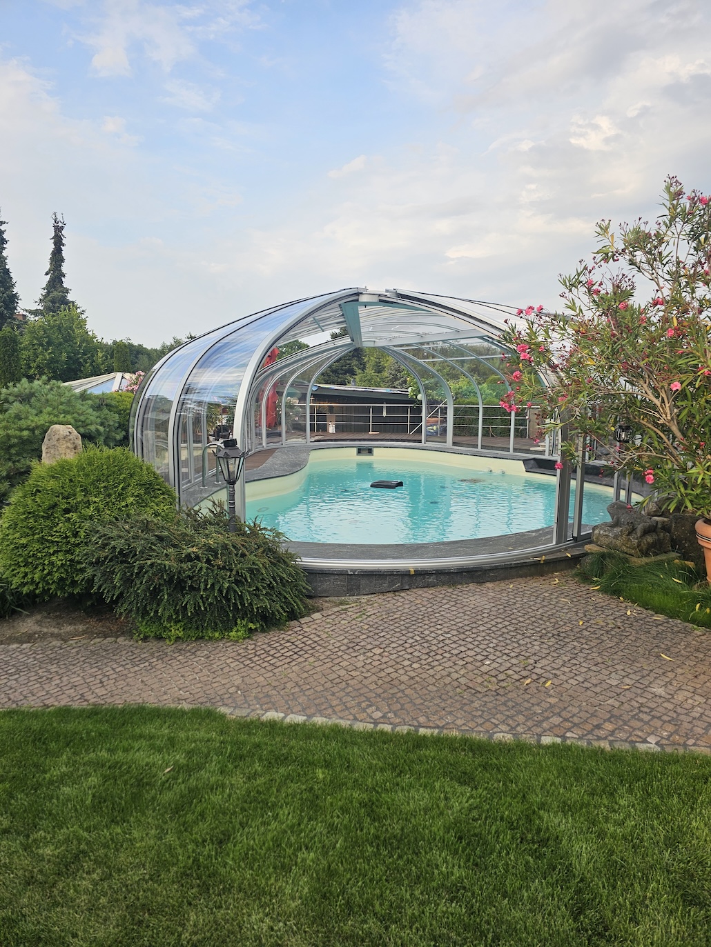 20260731_185222 Hohe transparente Poolüberdachung über einem geschwungenen Außenpool, umgeben von Sträuchern und Blumen