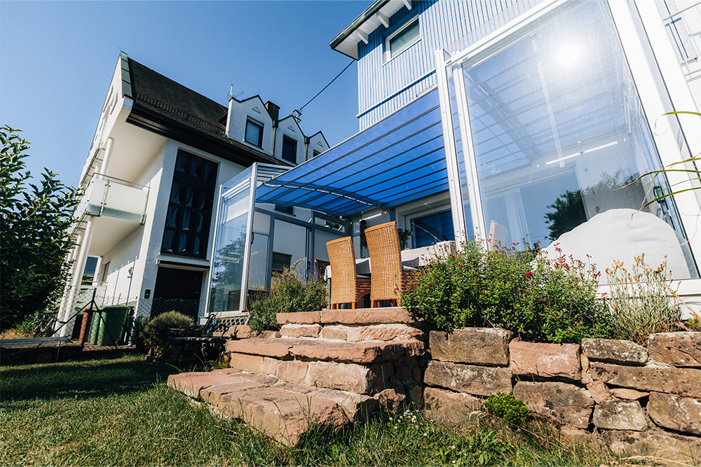 TOPAS Solar Veranda patio cover on the house, viewed from the garden, with stone steps and a planted patio edge.